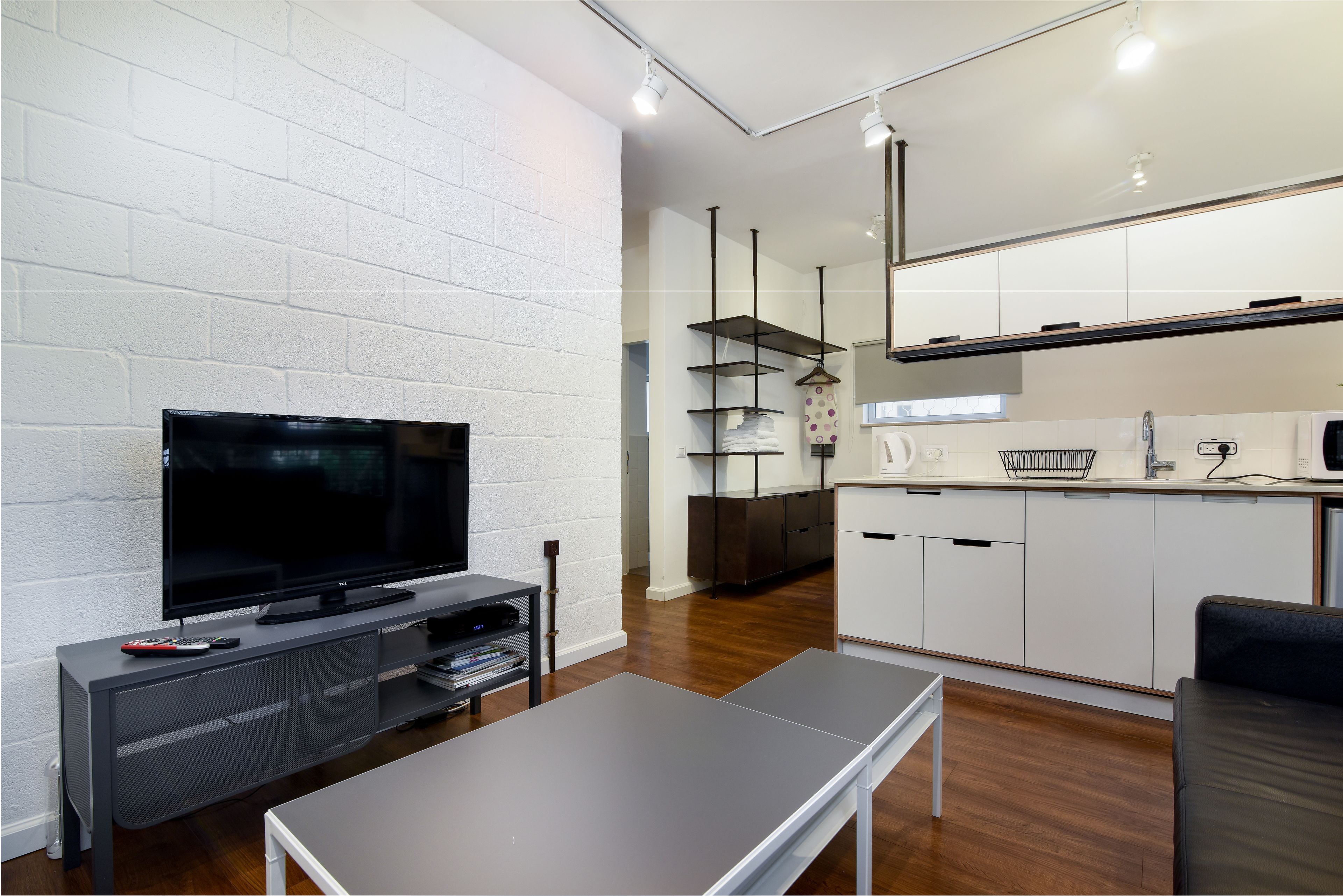 A modern living room with a flat-screen TV, a coffee table, and a sofa. There is a kitchen area with white cabinets and a microwave.