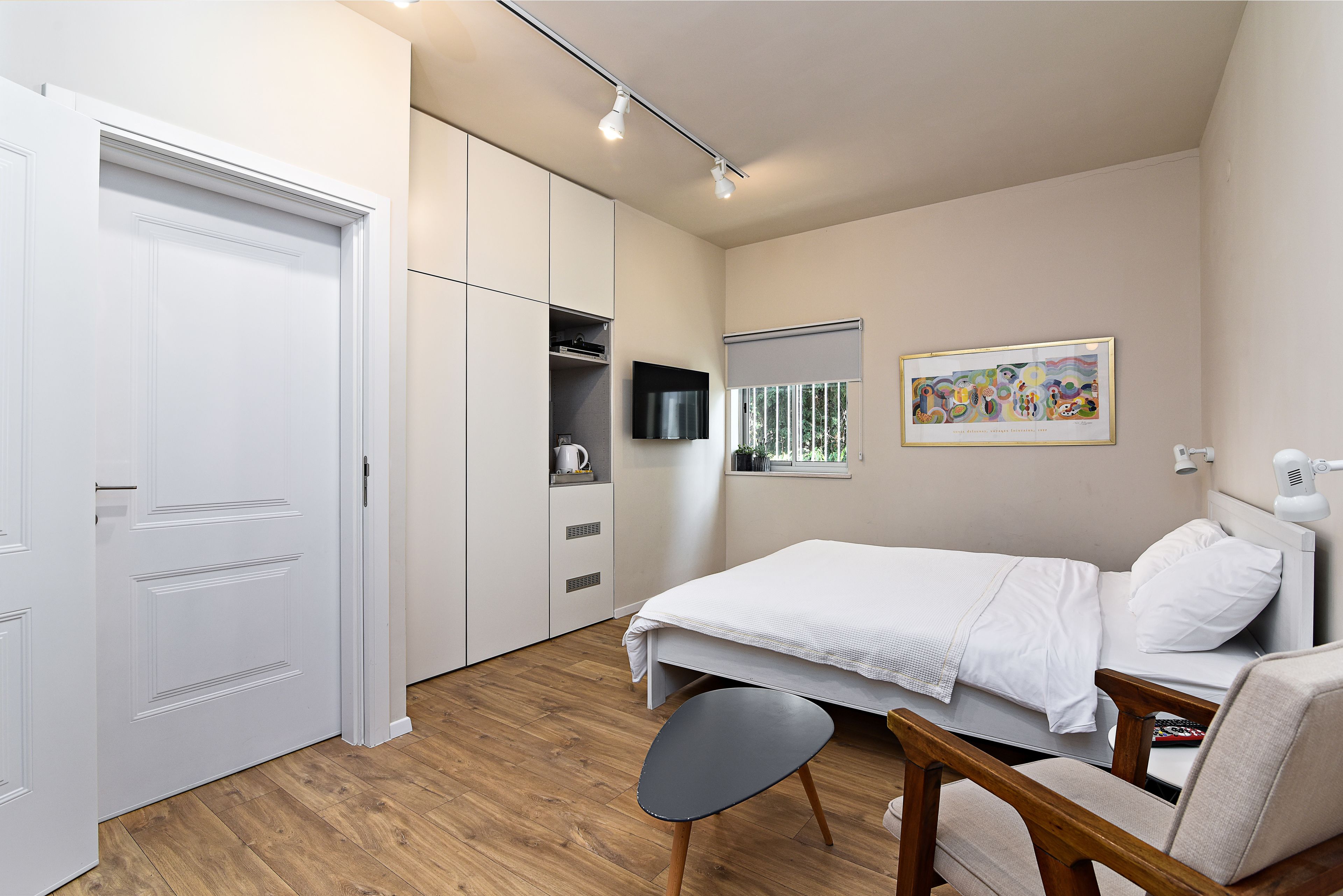 A bedroom with a bed, a chair, a desk, a wardrobe, and a television on the wall.