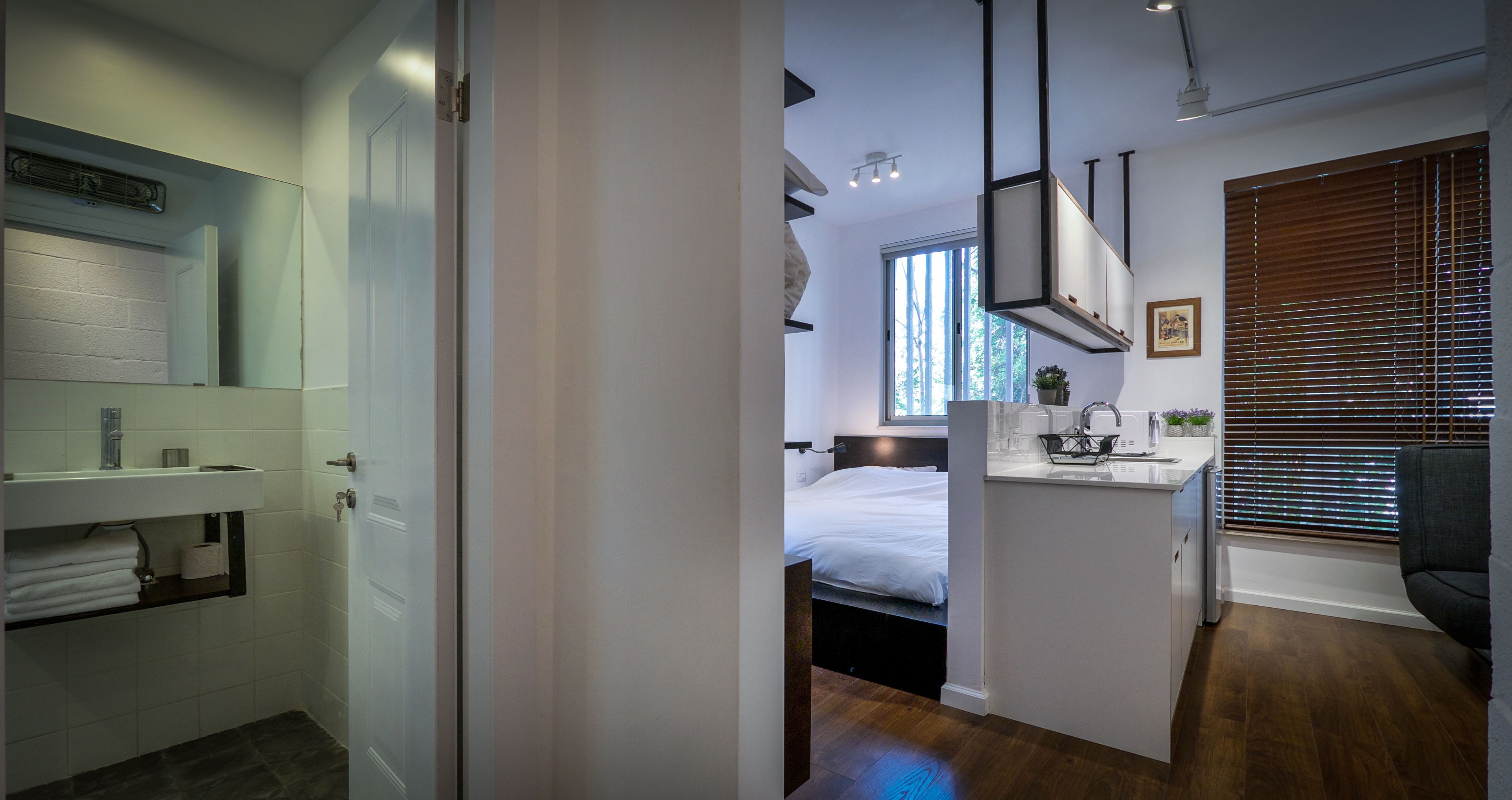 A modern hotel room with a large bed, a small kitchenette, a bathroom with a mirror and towel rack, and a window with blinds.