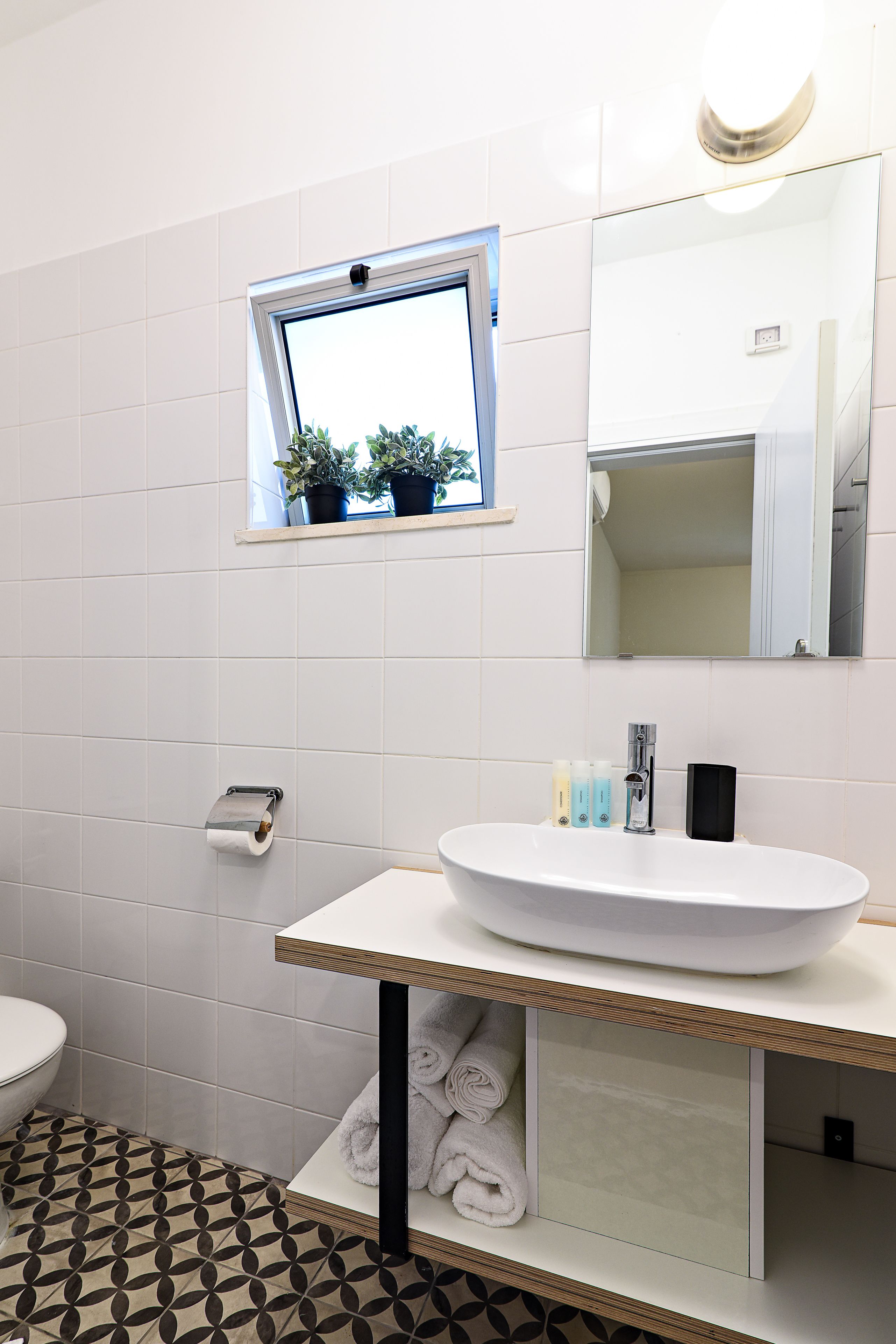 A bathroom with a white sink, a mirror, a window with plants, and a patterned floor.
