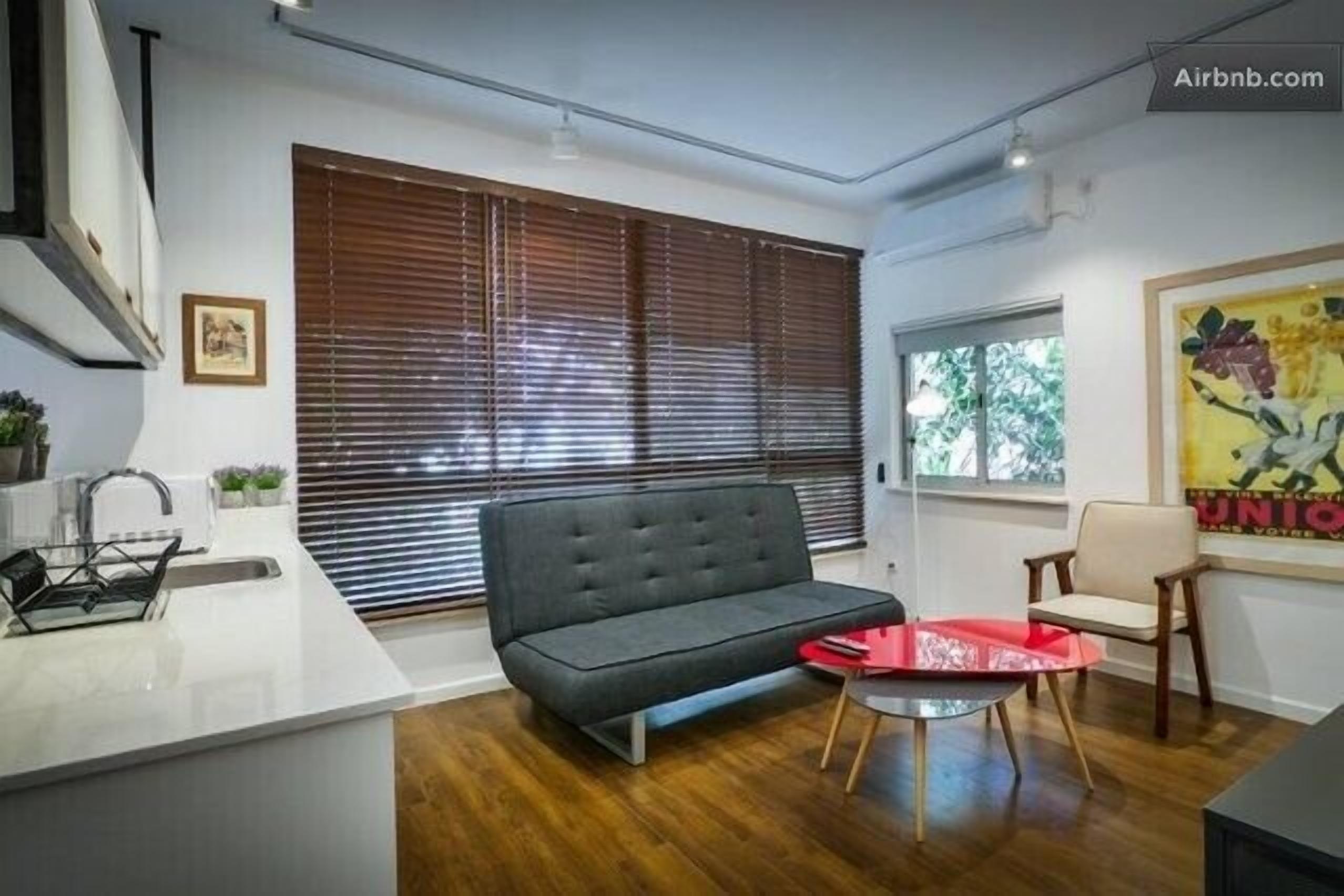 Una sala moderna con un sofá, una mesita y un sillón. Se ve una ventana con persianas, un lavamanos y una encimera.