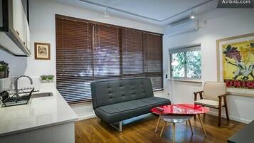 Una sala moderna con un sofá, una mesita y un sillón. Se ve una ventana con persianas, un lavamanos y una encimera.