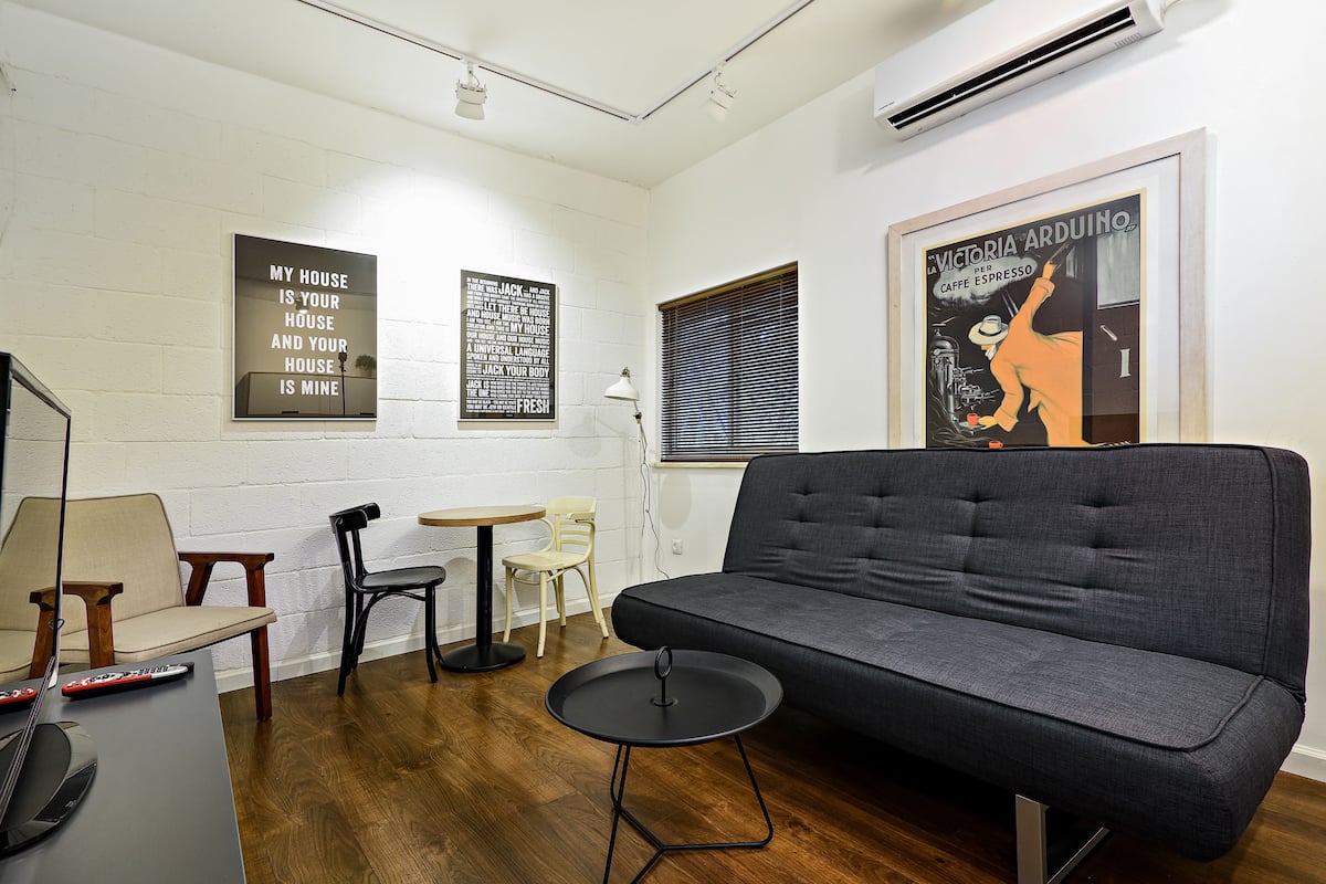 Un salon moderne avec un canapé gris, un plancher en bois et une petite table ronde. On y trouve deux chaises, une lampe et un tableau encadré accroché au mur.