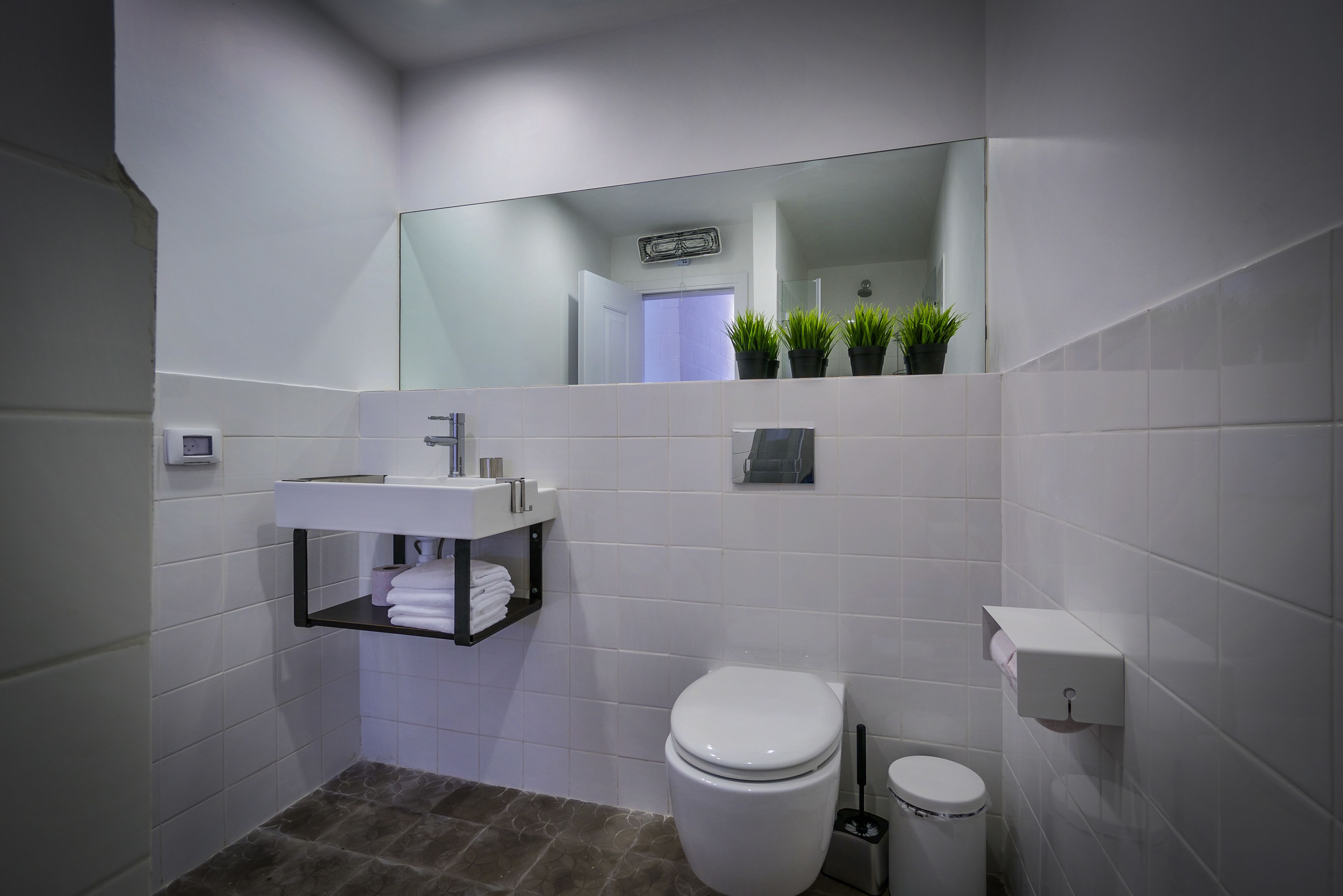 A modern bathroom with white tiles, a white toilet, a white sink with a towel rack, and a mirror above the sink.
