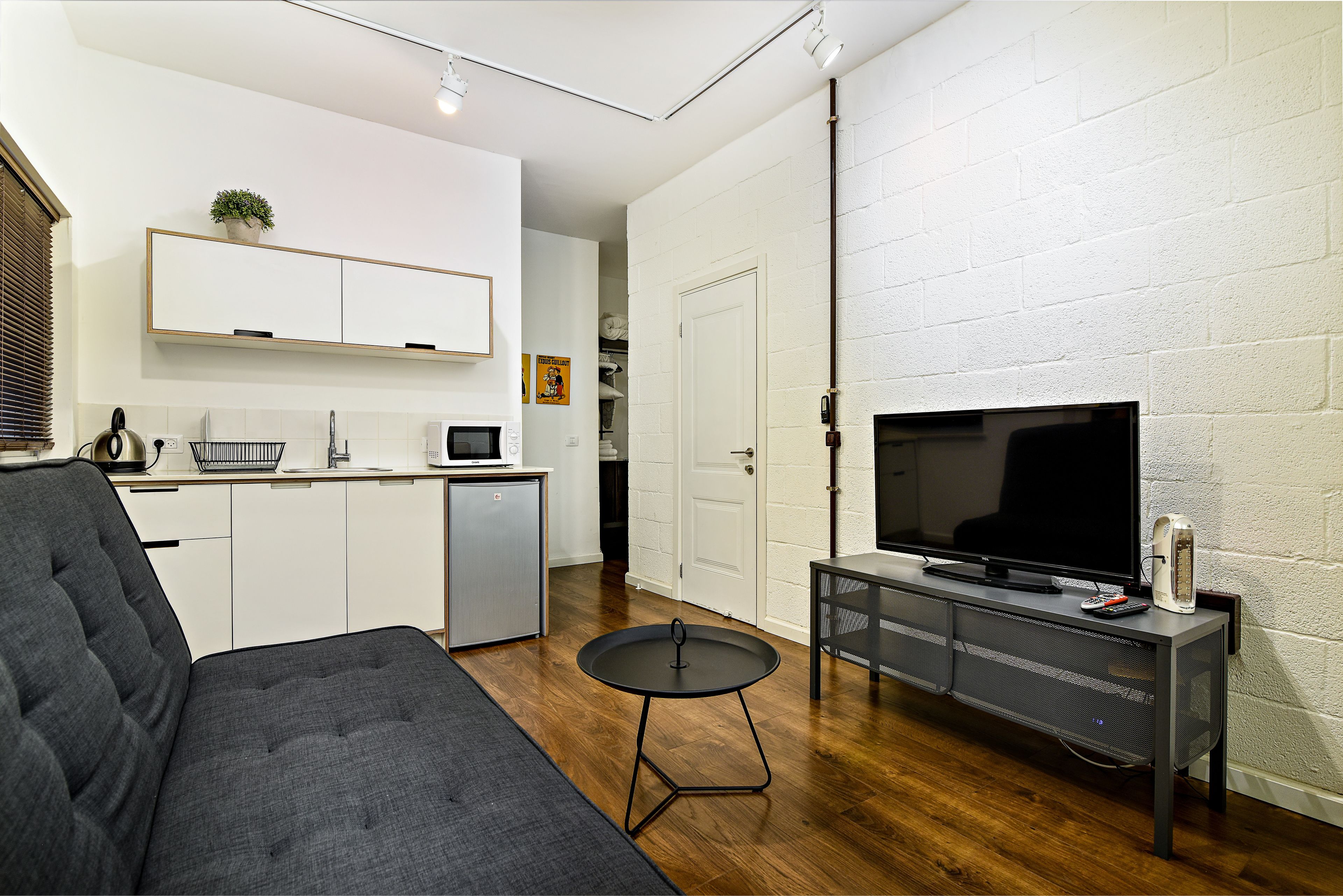 A compact living space with a grey sofa, a small round table, a TV on a stand, a kitchen area with a microwave, and a white brick wall.