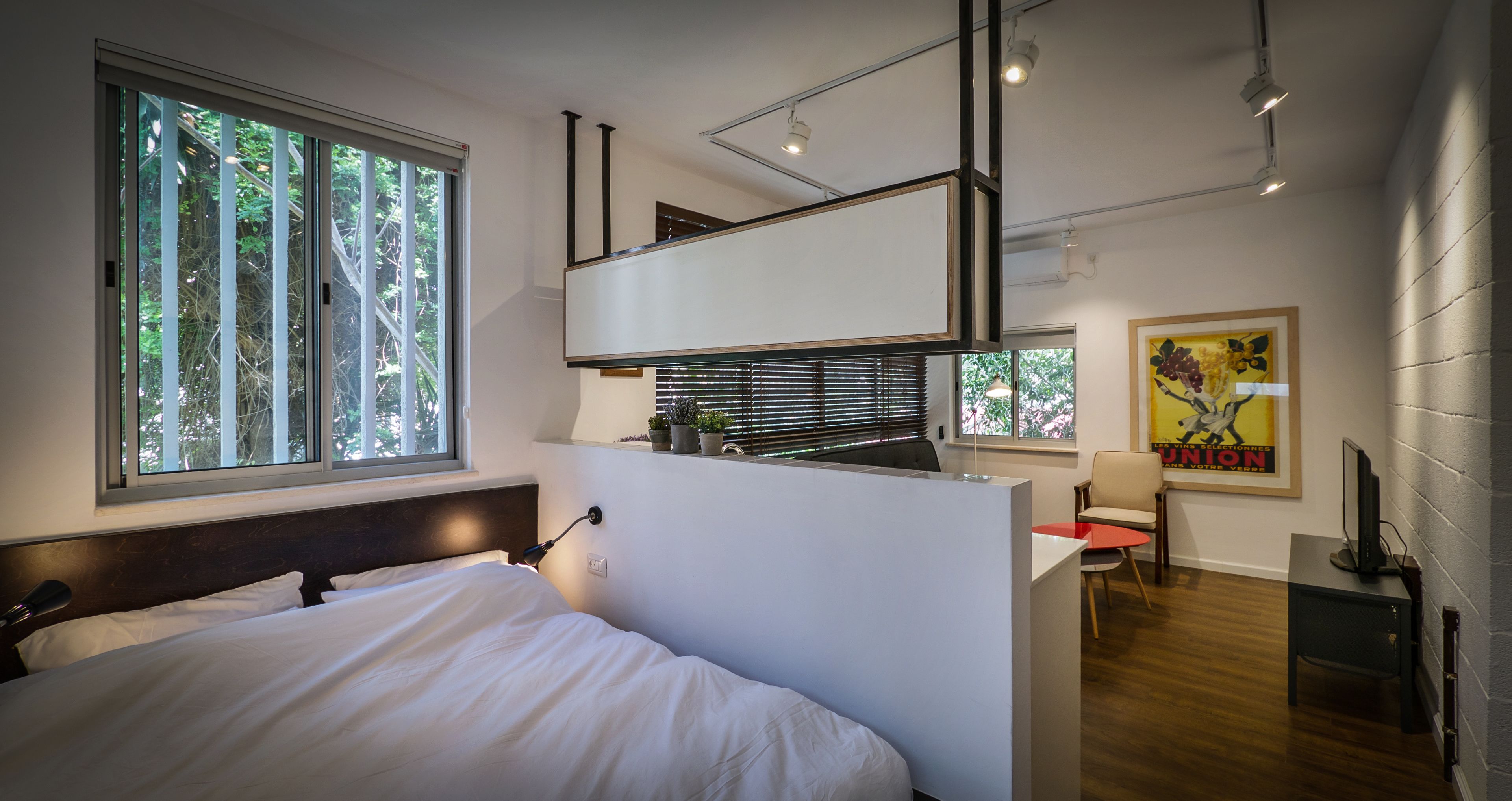 Un dormitorio moderno con una cama grande, un escritorio y una silla. Hay una ventana con persianas, una lámpara de techo y un televisor montado en la pared.