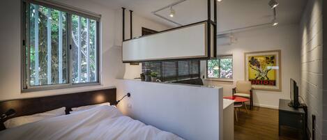 Un dormitorio moderno con una cama grande, un escritorio y una silla. Hay una ventana con persianas, una lámpara de techo y un televisor montado en la pared.