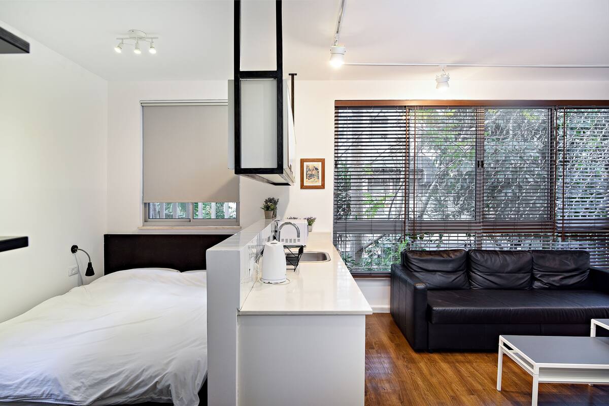 Une chambre moderne équipée d’un lit, d’un coin cuisine, d’un canapé en cuir noir et d’une grande fenêtre avec des stores.