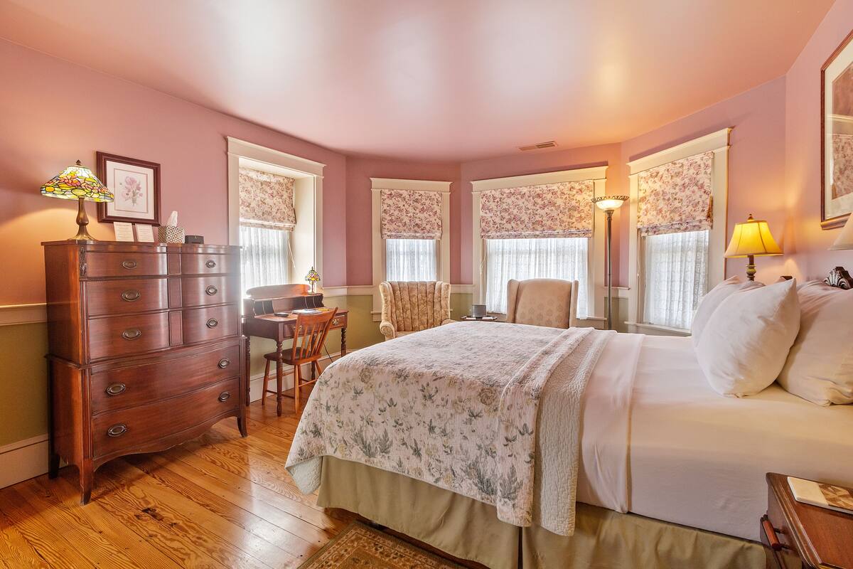 A bedroom with a wooden dresser, a bed with a floral bedspread, a chair, and a desk with a lamp.