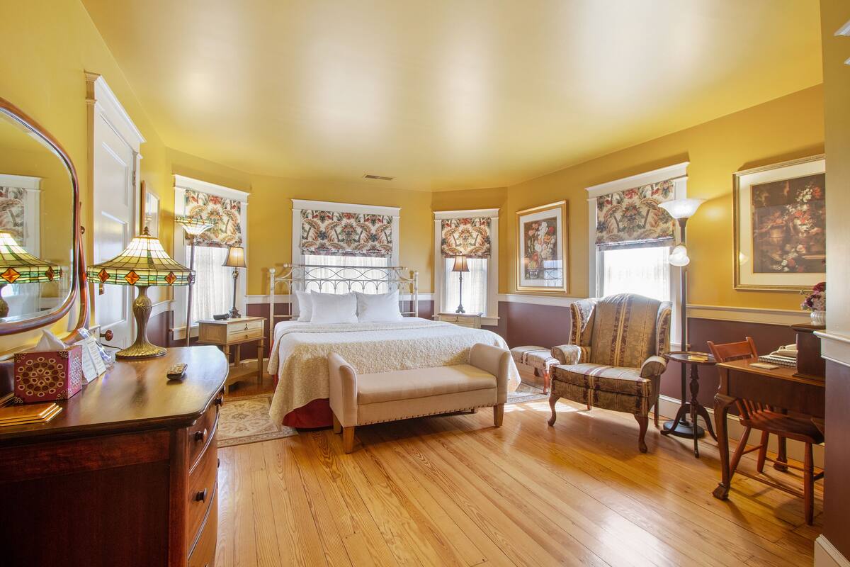 A well-lit bedroom with a large bed, a sitting area with a chair and table, and a dresser with a mirror and lamp.
