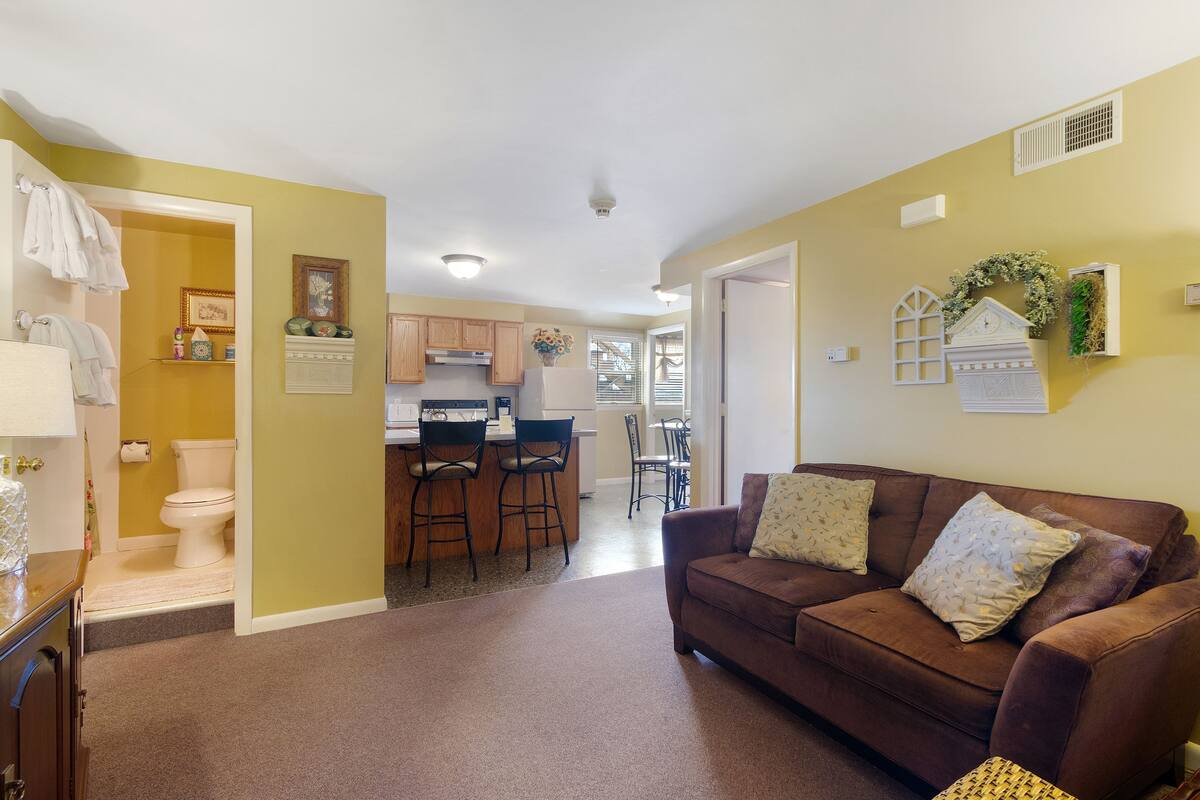A living room with a brown sofa, a kitchen in the background, and a bathroom on the left.