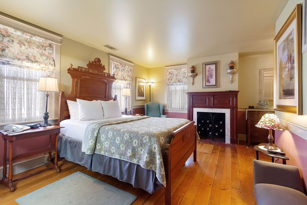 A traditional bedroom with a wooden bed, a desk with a lamp, a chair, a fireplace, and a framed picture on the wall.