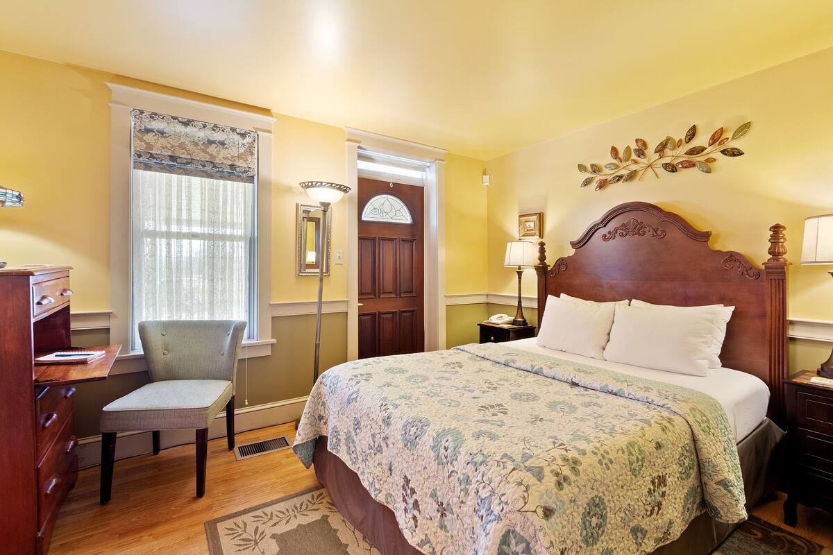 A bedroom with a large bed, a chair, a desk, and a window with curtains.