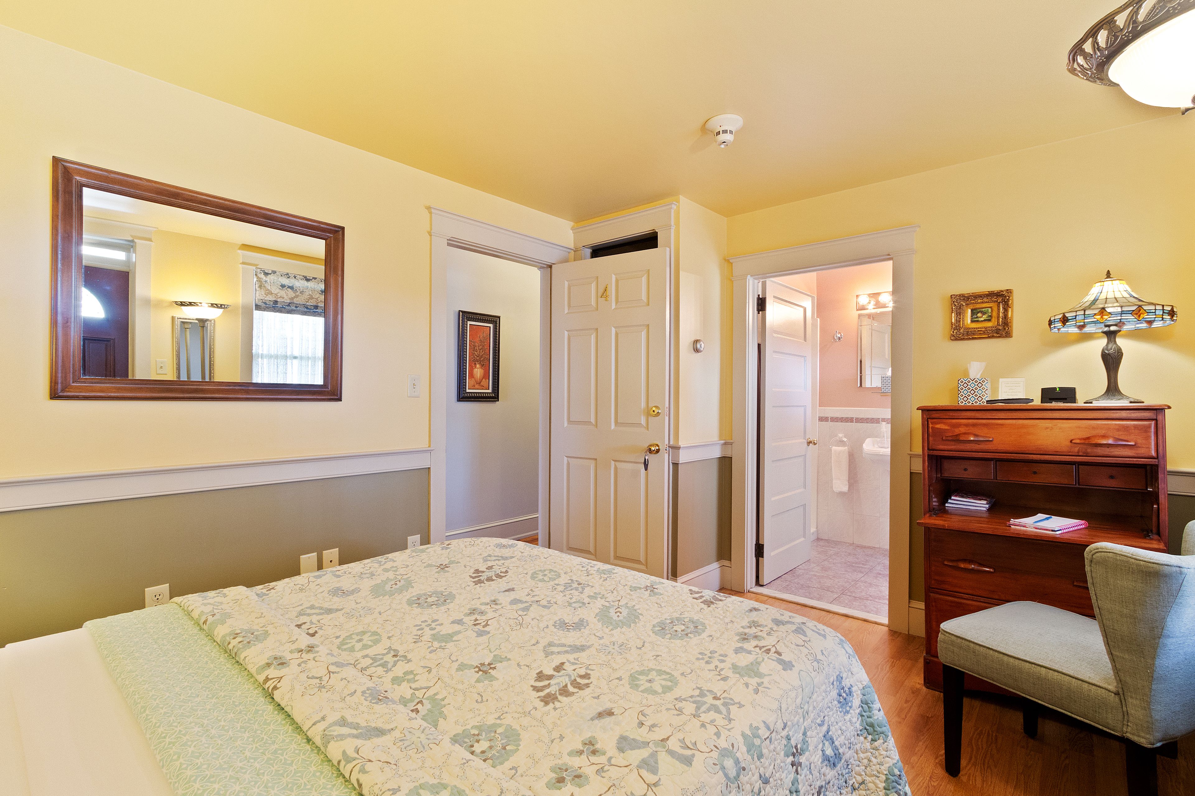 A bedroom with a bed, a dresser, a chair, a mirror, and a door leading to another room.