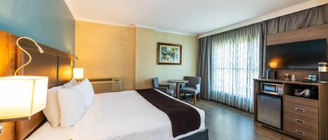 Quarto de hotel com uma cama grande, uma escrivaninha com luminária, uma televisão, uma mesinha e um micro-ondas.