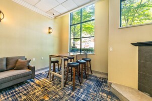 Una habitación con sofá, una mesa con taburetes, alfombra estampada, chimenea y ventana con vistas a árboles.