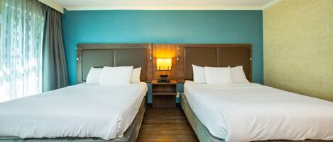 A hotel room with two beds, a wooden headboard, a nightstand with a lamp, and a window with curtains.