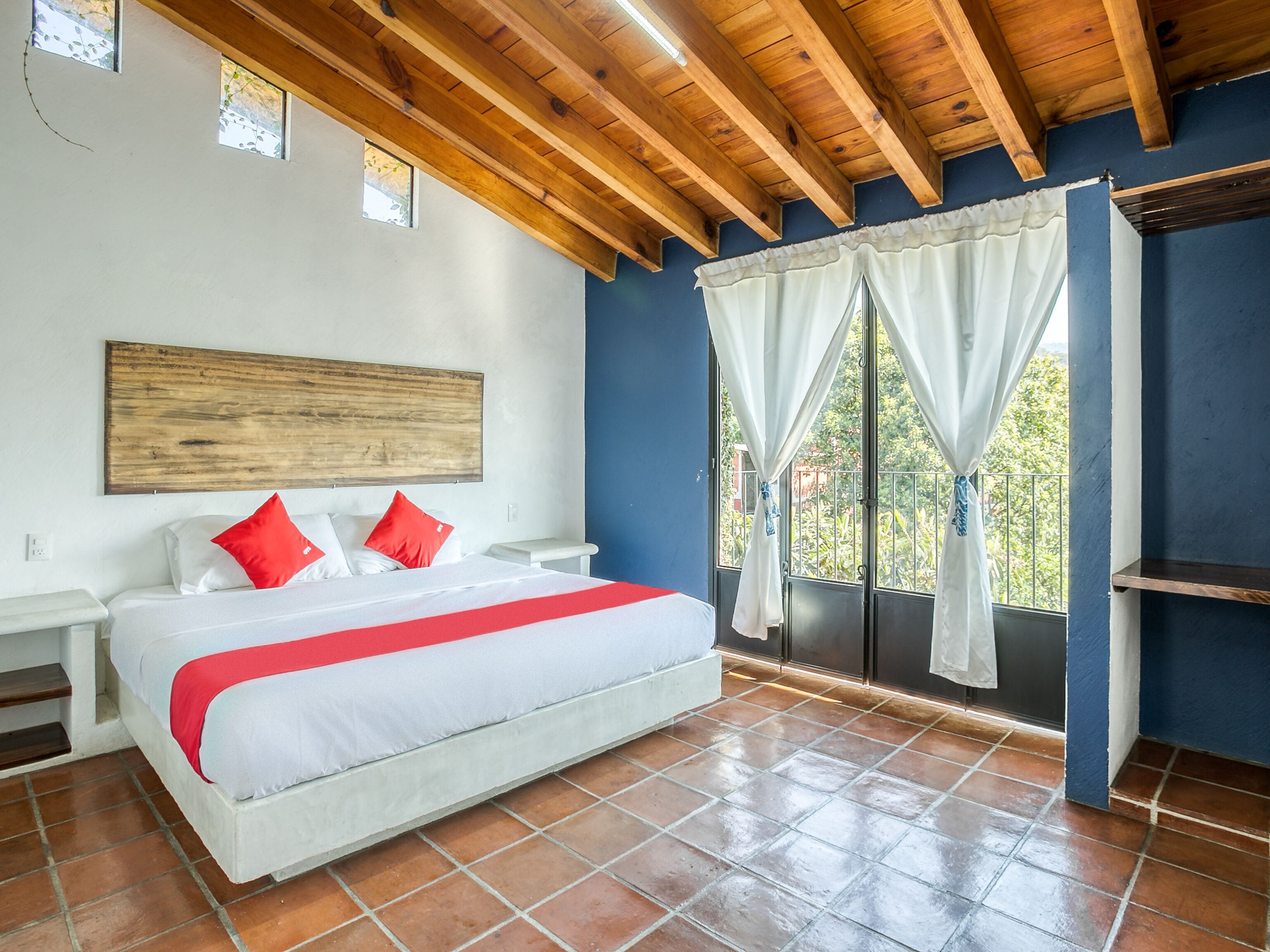 A bedroom with a wooden ceiling, a bed with white bedding and red pillows, a large window with white curtains, and a blue wall.