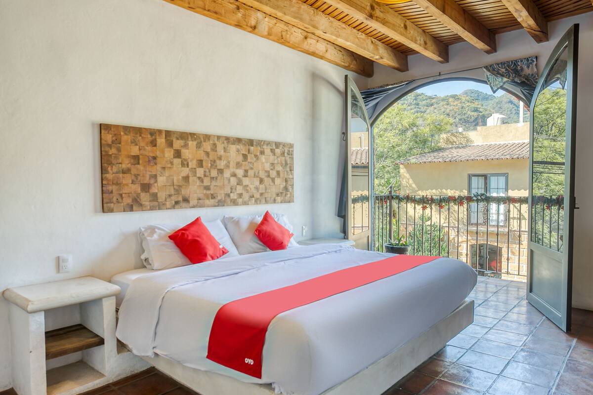 Ein Schlafzimmer mit einem großen Bett, einem Holz-Kopfbrett, einem Nachttisch und Blick auf einen Balkon mit Geländer und Pflanzen in Töpfen.