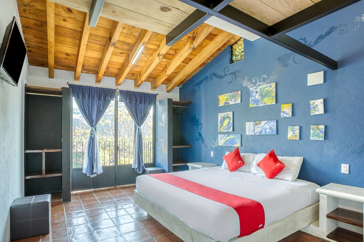 Ein Schlafzimmer mit Holzbalkendecke, einem Bett mit rot-weiß gestreifter Bettwäsche, einem Fenster mit Vorhängen und einem Bild an der Wand.