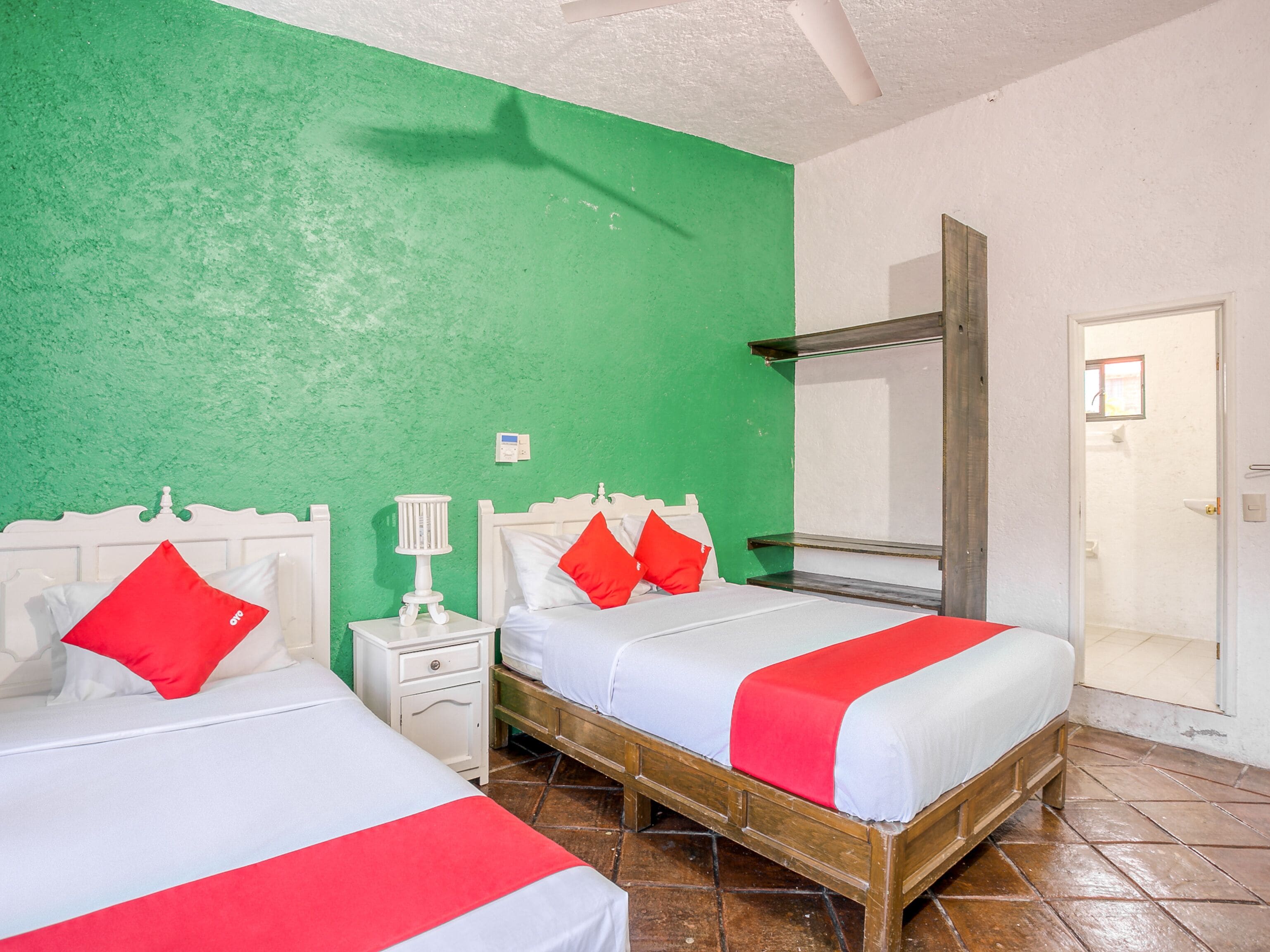 Una habitación con dos camas, cada una con ropa de cama blanca y detalles en rojo, una mesita de noche pequeña con lámpara y un espejo sobre las camas.