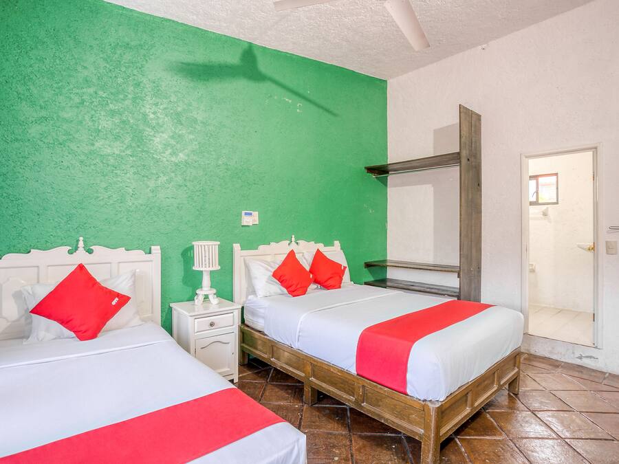 A room with two beds, each with white bedding and red accents, a small bedside table with a lamp, and a mirror above the beds.