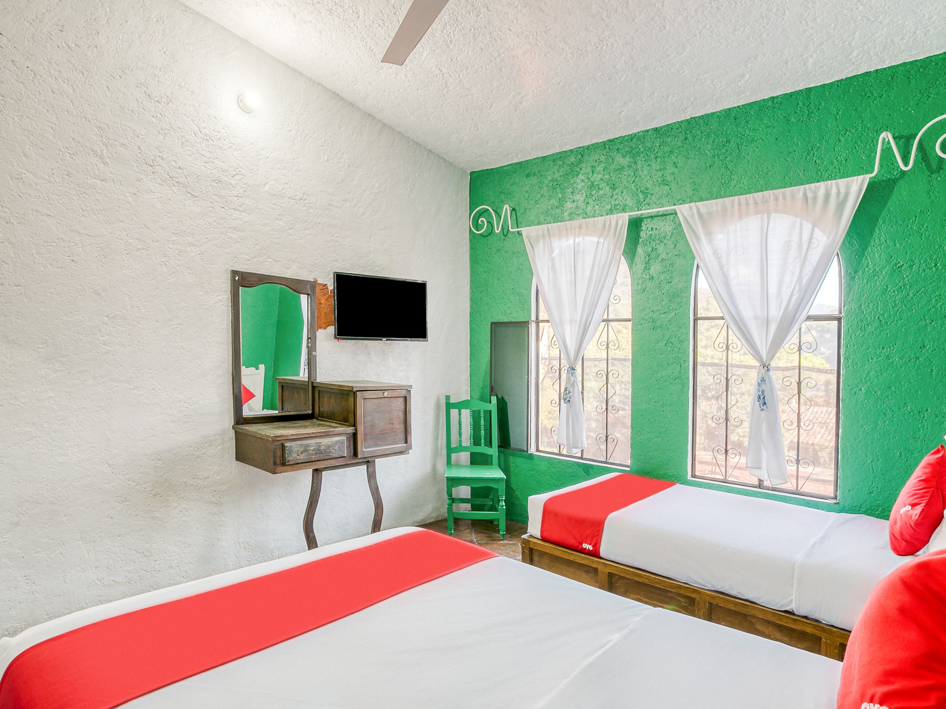 A room with two beds, a small desk, a mirror, a television, and a window with curtains.