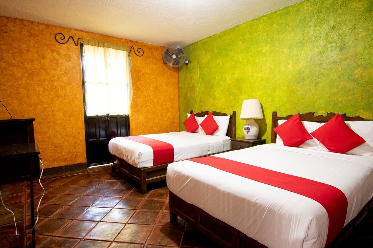 Ein Hotelzimmer mit zwei Betten, einem Ventilator, einer Lampe und einem Fenster mit Vorhängen.