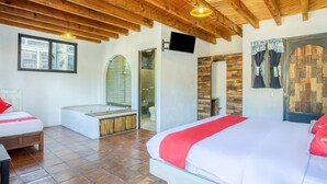 Habitación de hotel con techo de madera, una cama grande con ropa de cama roja y blanca, una mesita pequeña, un banco, una bañera y zona de ducha.