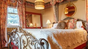 Ein Schlafzimmer mit einem großen Bett, einem verzierten Metall-Kopfteil, blumigen Vorhängen, einem Spiegel und einer Kandelaber.
