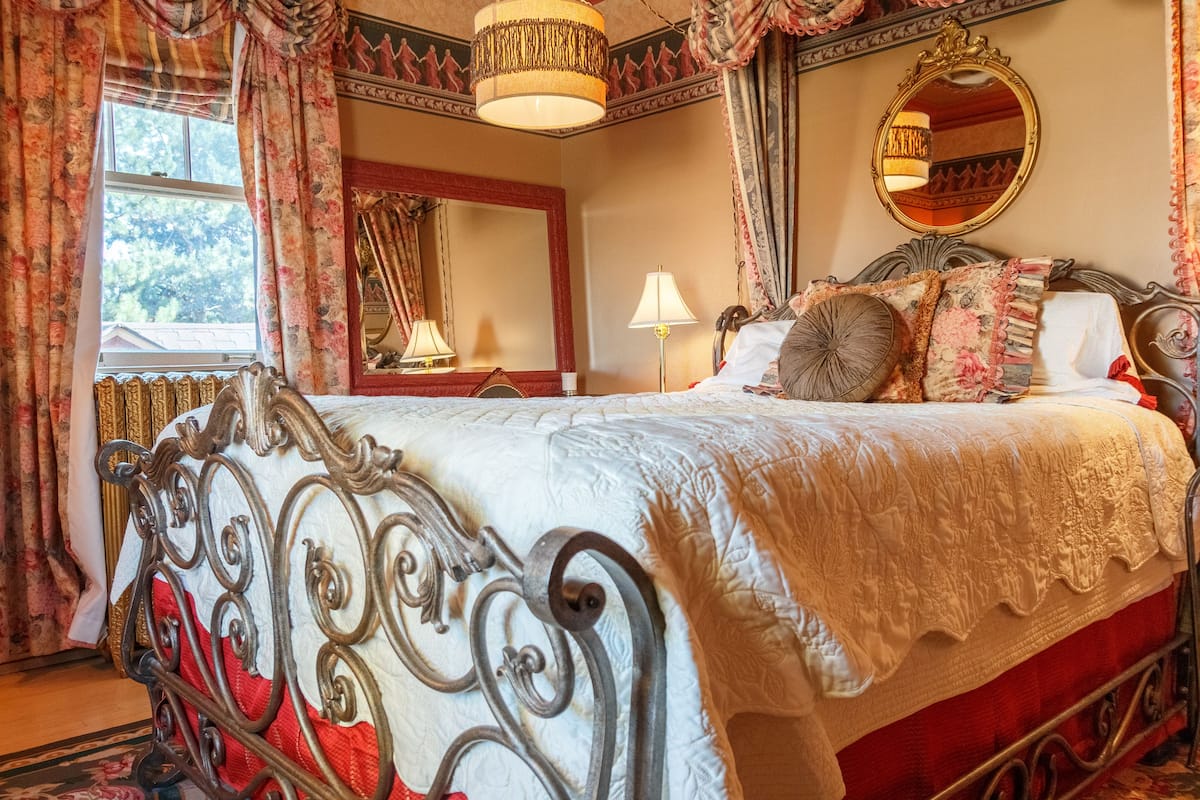 A bedroom with a large bed, ornate metal headboard, floral curtains, a mirror, and a chandelier.