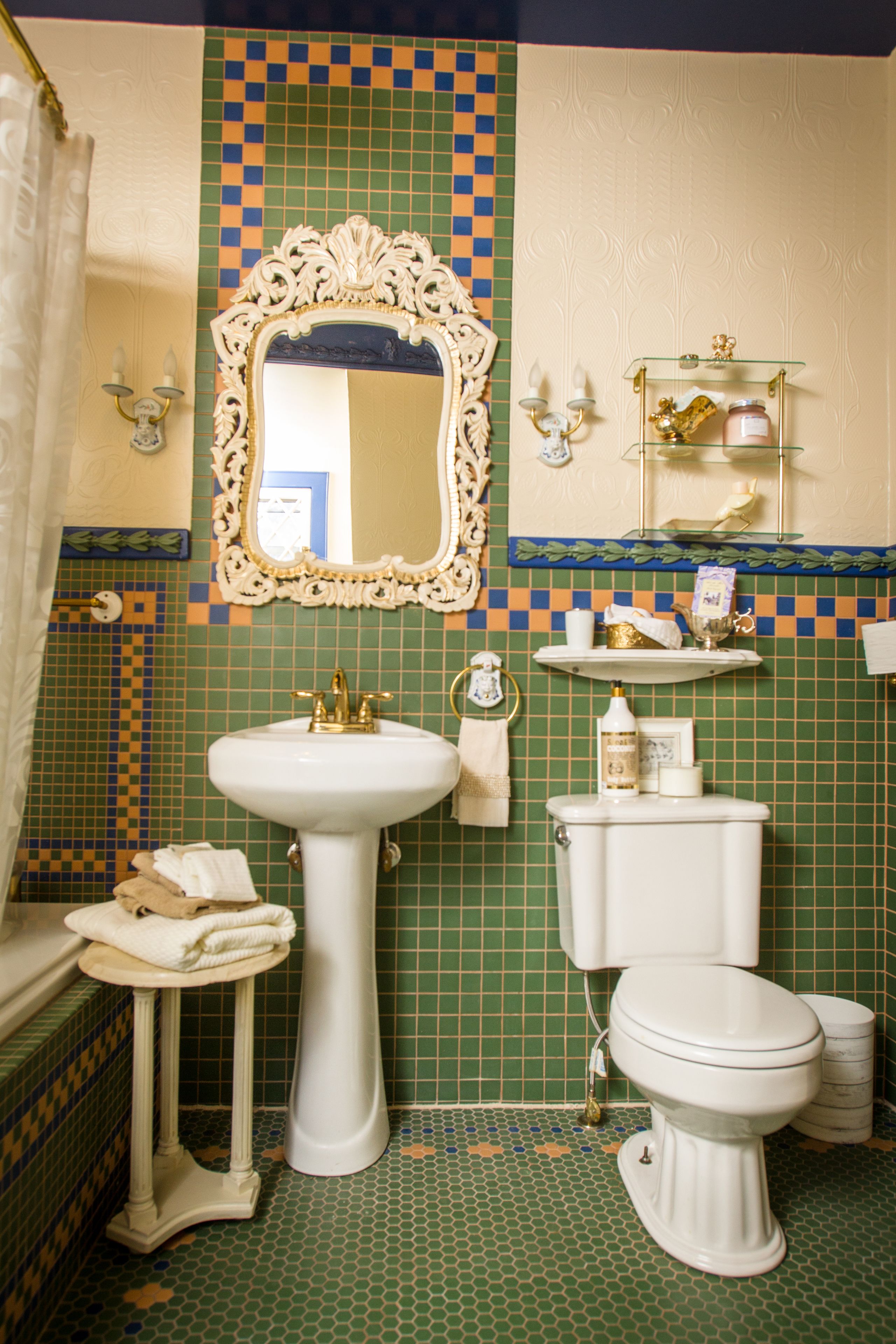Ein Badezimmer mit einem Standwaschbecken, einer Toilette, einem Spiegel mit aufwendigem Rahmen und einem Ablagefach mit Pflegeprodukten.