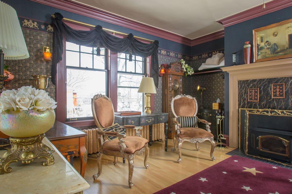 A room with two ornate chairs, a fireplace, and a large vase with flowers on a table.