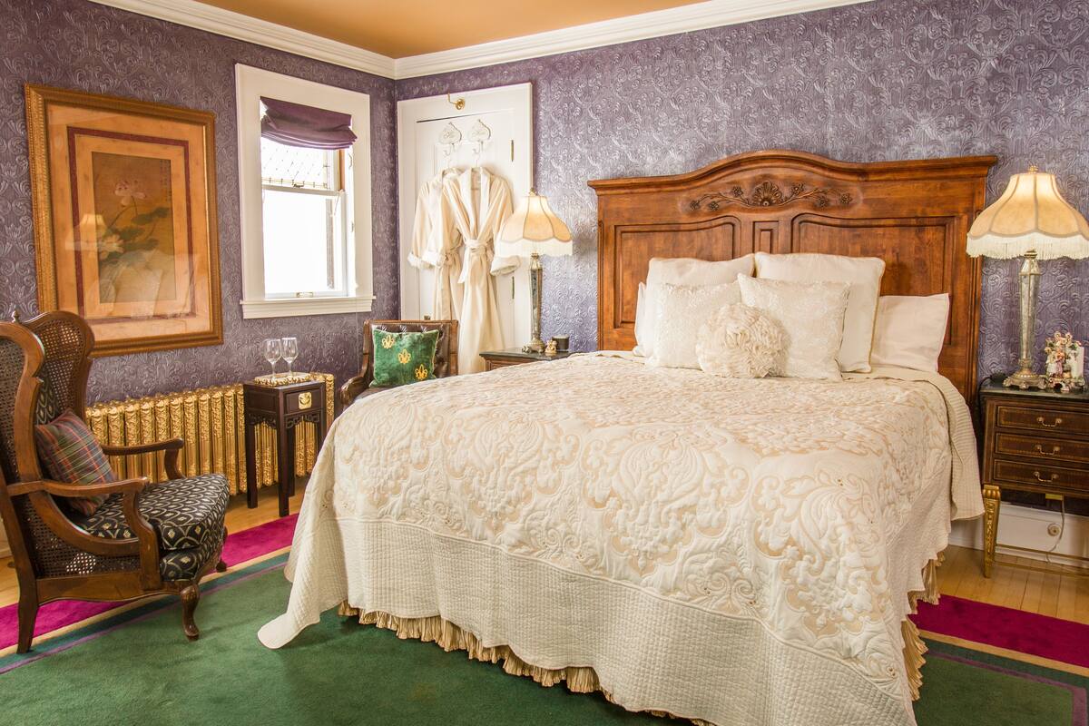 A bedroom with a large bed, a wooden headboard, a green carpet, a wooden chair, a framed picture, and a window with a purple curtain.