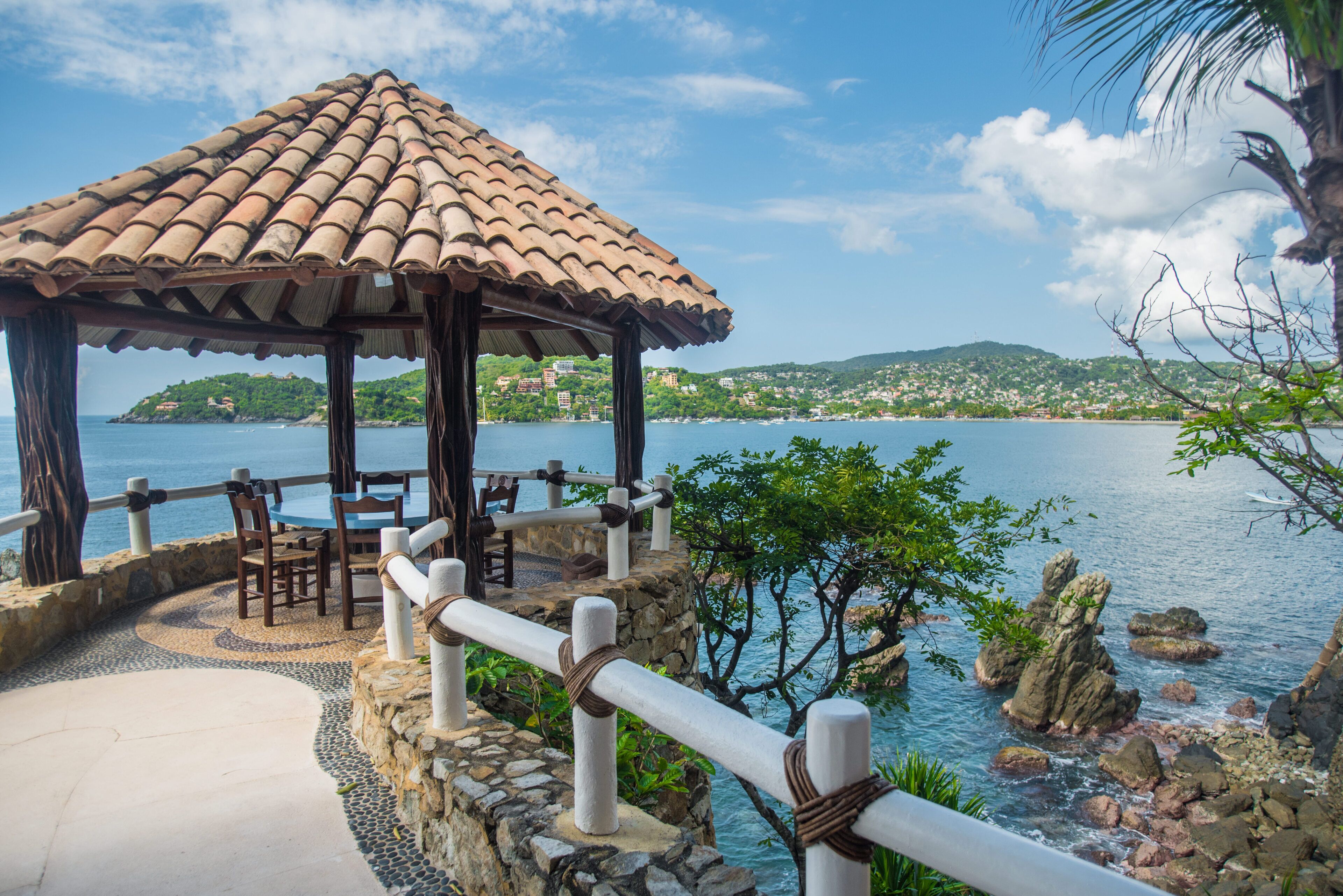 A gazebo with a tiled roof overlooking a coastal area with a view of the sea and hills.