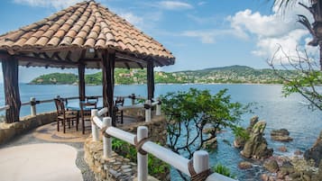 A gazebo with a tiled roof overlooking a coastal area with a view of the sea and hills.