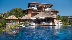 Un resort con una amplia piscina, estructuras de techo de paja y un edificio de varios pisos con balcones.