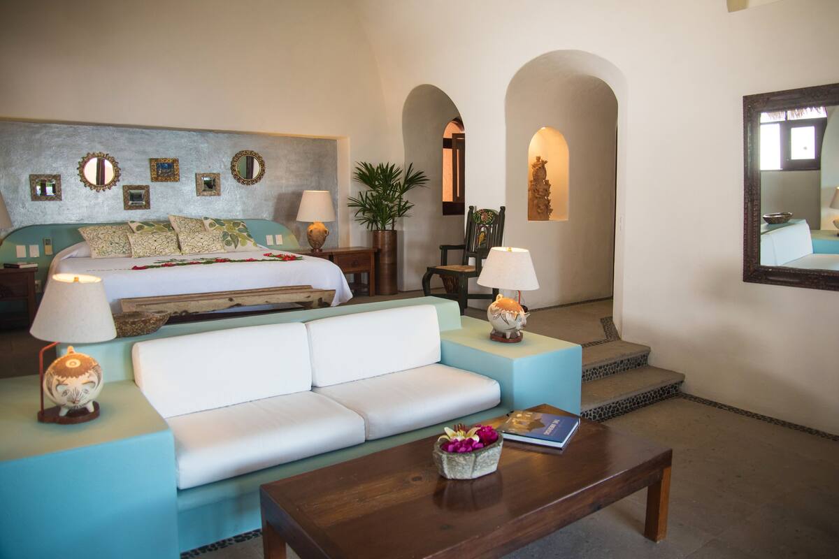 A hotel room with a bed, a white sofa, a wooden coffee table, and a small table with a lamp.