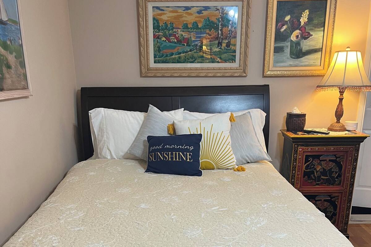 Ein ordentlich gemachtes Bett mit einer quiltschönen Bettdecke mit floralem Muster, einem Kissen mit der Aufschrift 'good morning SUNSHINE' und zwei gerahmten Bildern an der Wand.