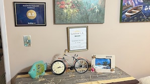 A wall with framed awards, a painting of a garden, a welcome sign, a bicycle, and a small fish figurine. - Sunshine LA BnB (Van Nuys)