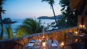 An outdoor dining area with a stone wall, wooden table, and chairs, overlooking a beach and ocean. - Villa El Ensueño by La Casa Que Canta (Zihuatanejo)