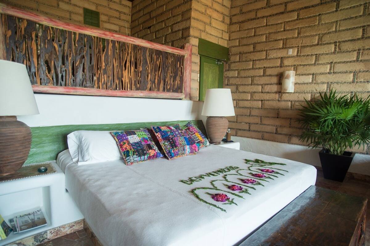 Ein Schlafzimmer mit Ziegelwand, einem grünen Kopfteil, einem weißen Bett mit Stickereien, farbenfrohen Kissen, zwei Nachttischlampen und einem Blumentopf.