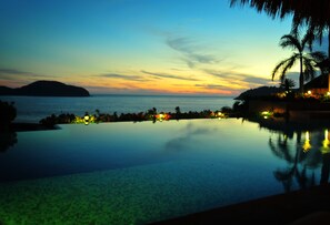 A poolside view with a sunset over the water, palm trees, and a distant island.