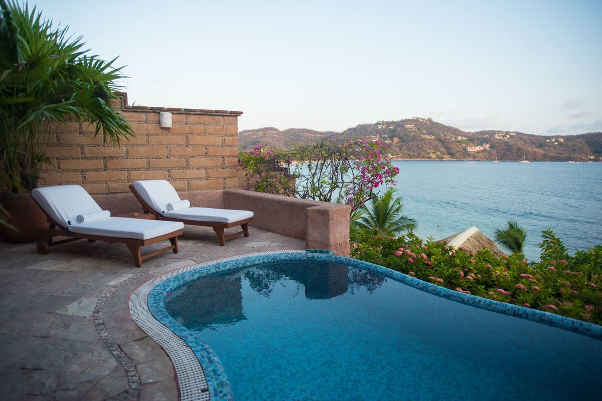 Ein Pool mit zwei Liegestühlen, einer Ziegelwand und Blick auf das Meer.
