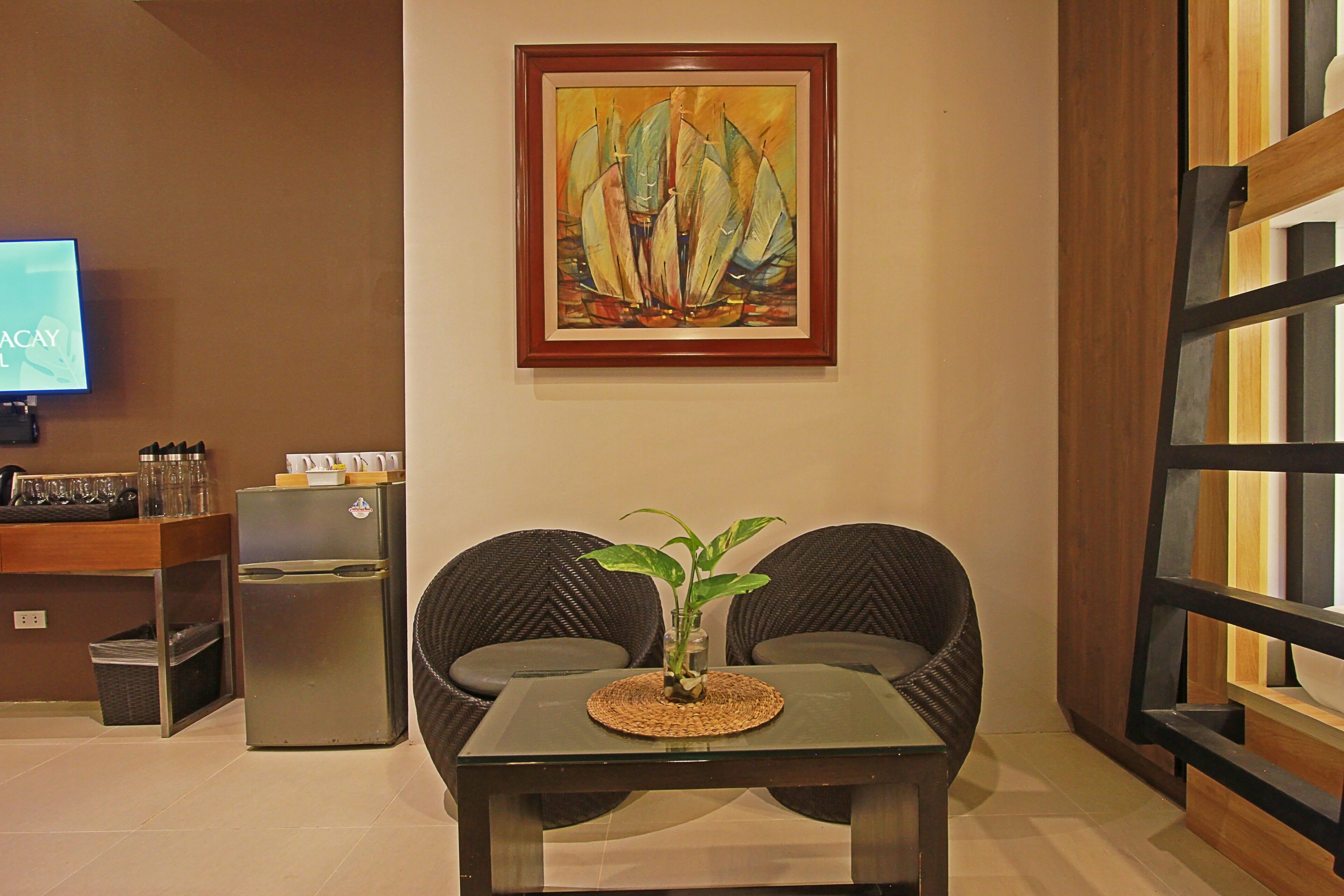 A room with a television, a refrigerator, two wicker chairs, a small table with a plant, and a painting on the wall.
