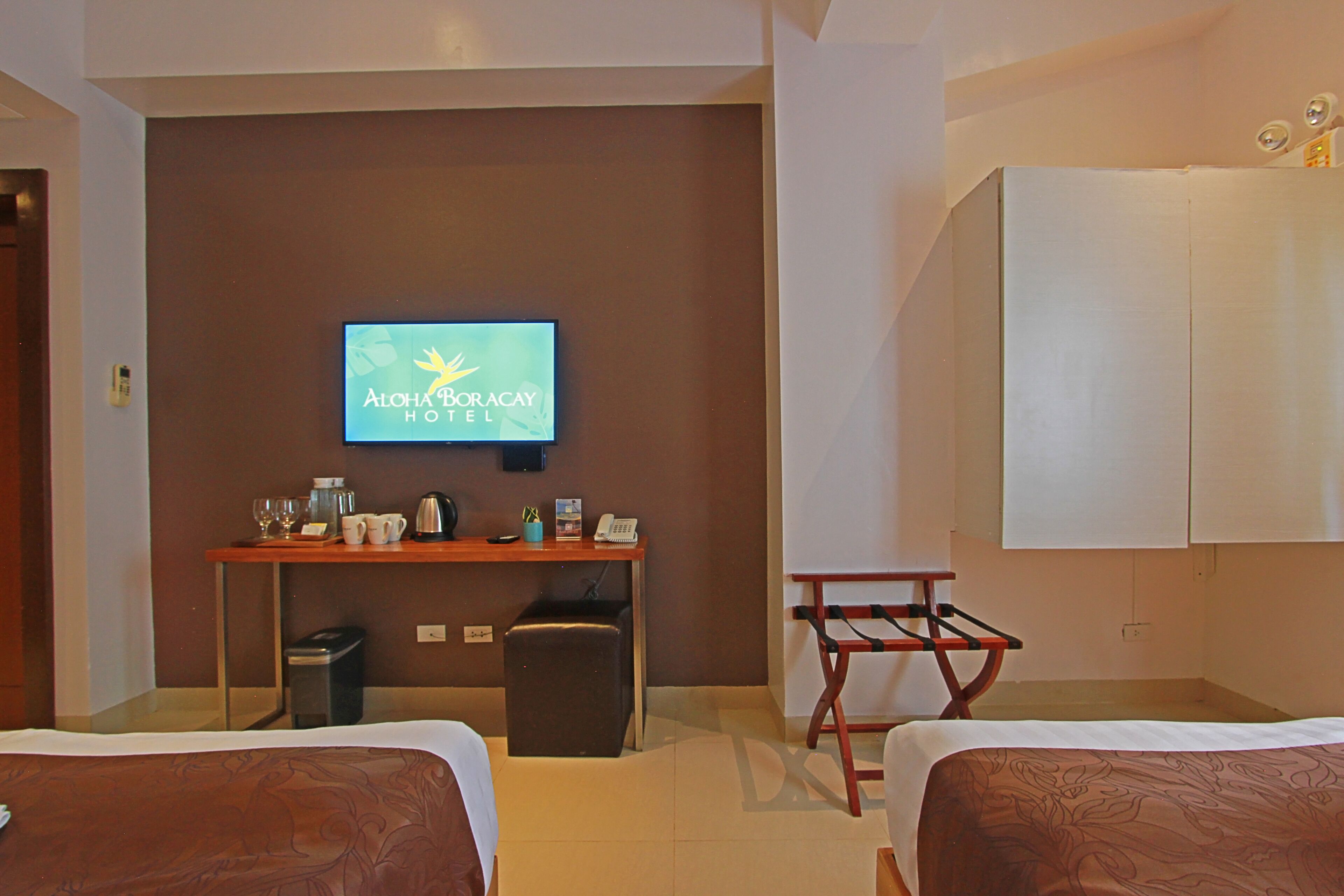 A hotel room with two beds, a desk with a television displaying the hotel name, a small trash bin, and a coat rack.