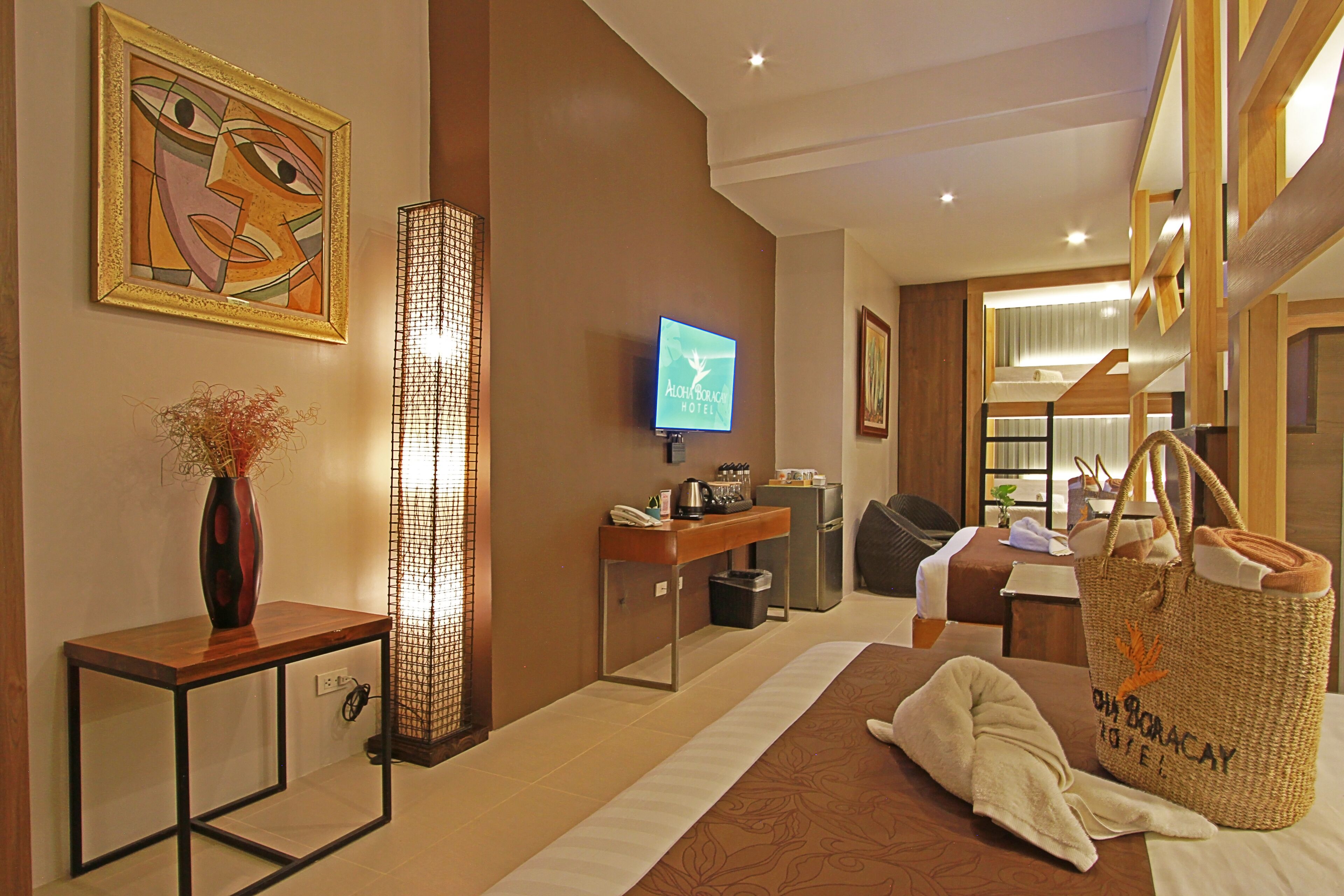 Una habitación de hotel con una cama, un escritorio, un televisor, un bolso tejido grande y un cuadro en la pared.