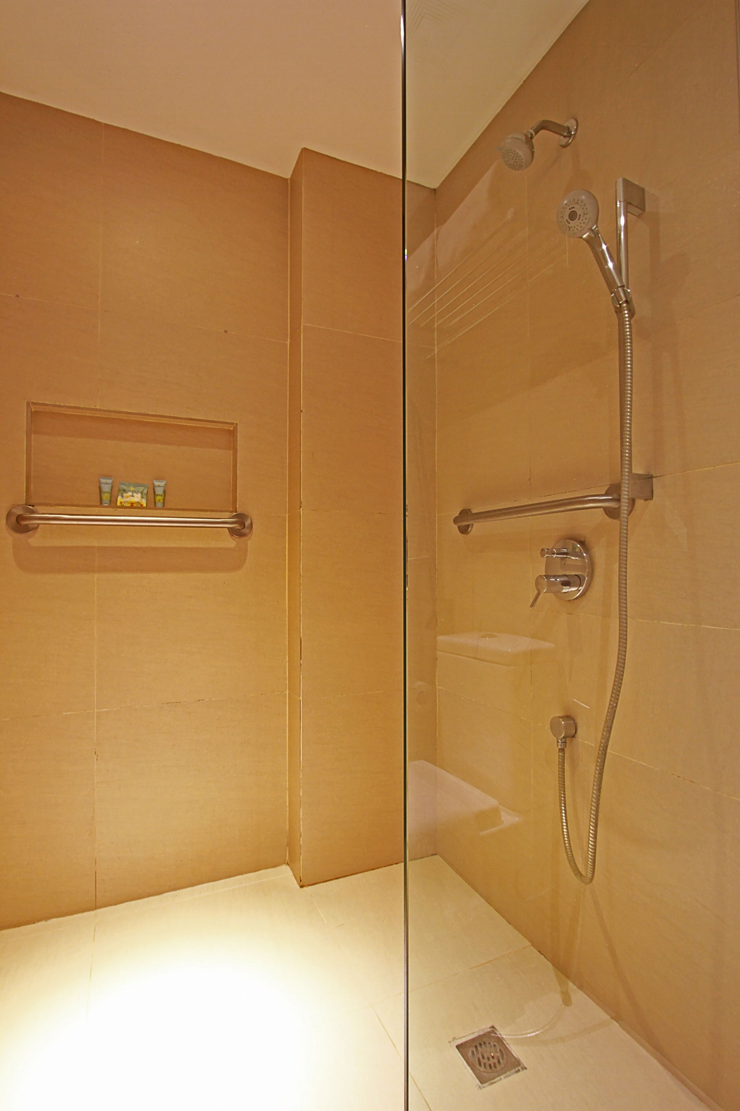 A glass-enclosed shower area with a handheld showerhead, a wall-mounted faucet, and a shelf with toiletries.