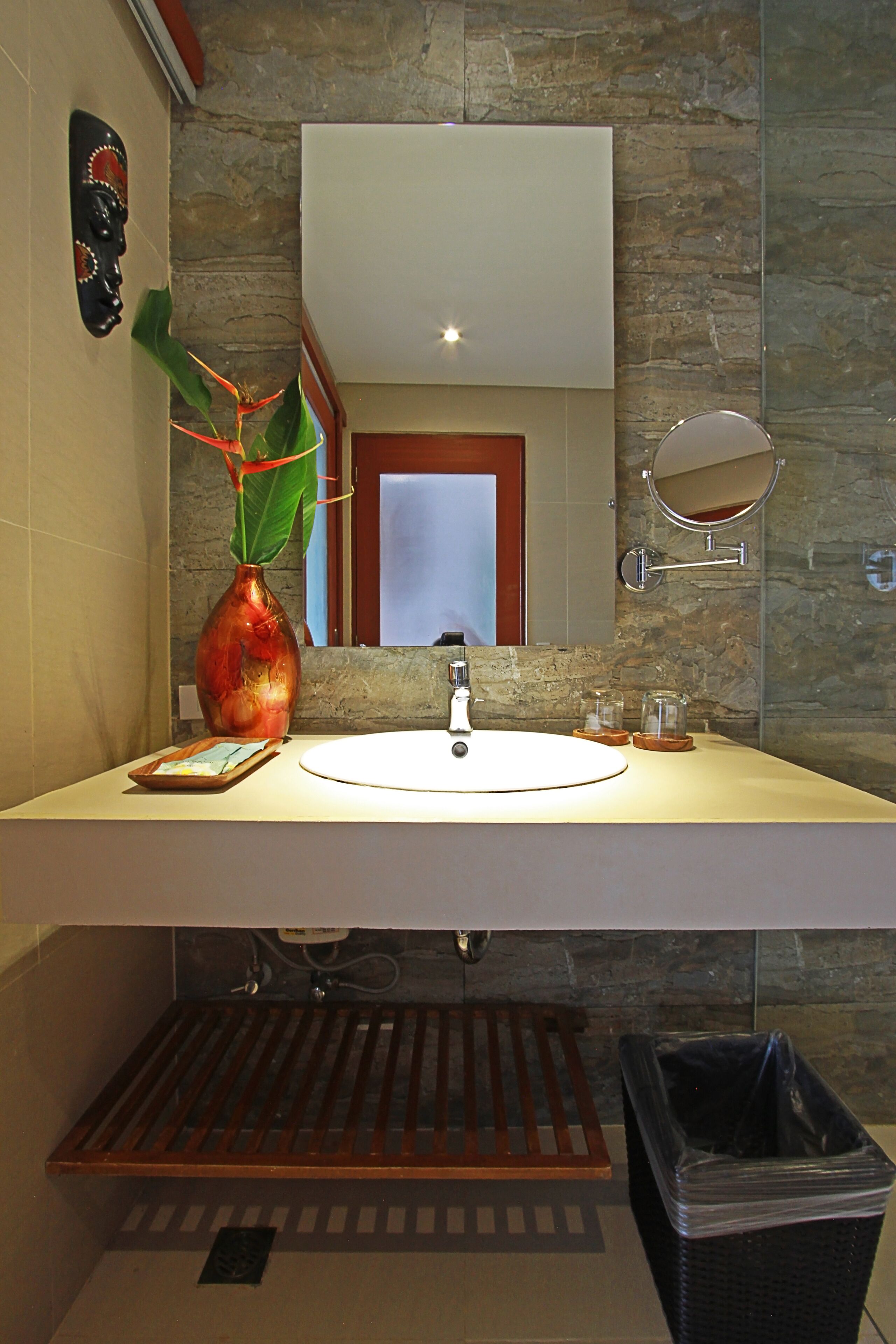 A bathroom with a white sink, a mirror, a vase with a plant, and a trash bin.
