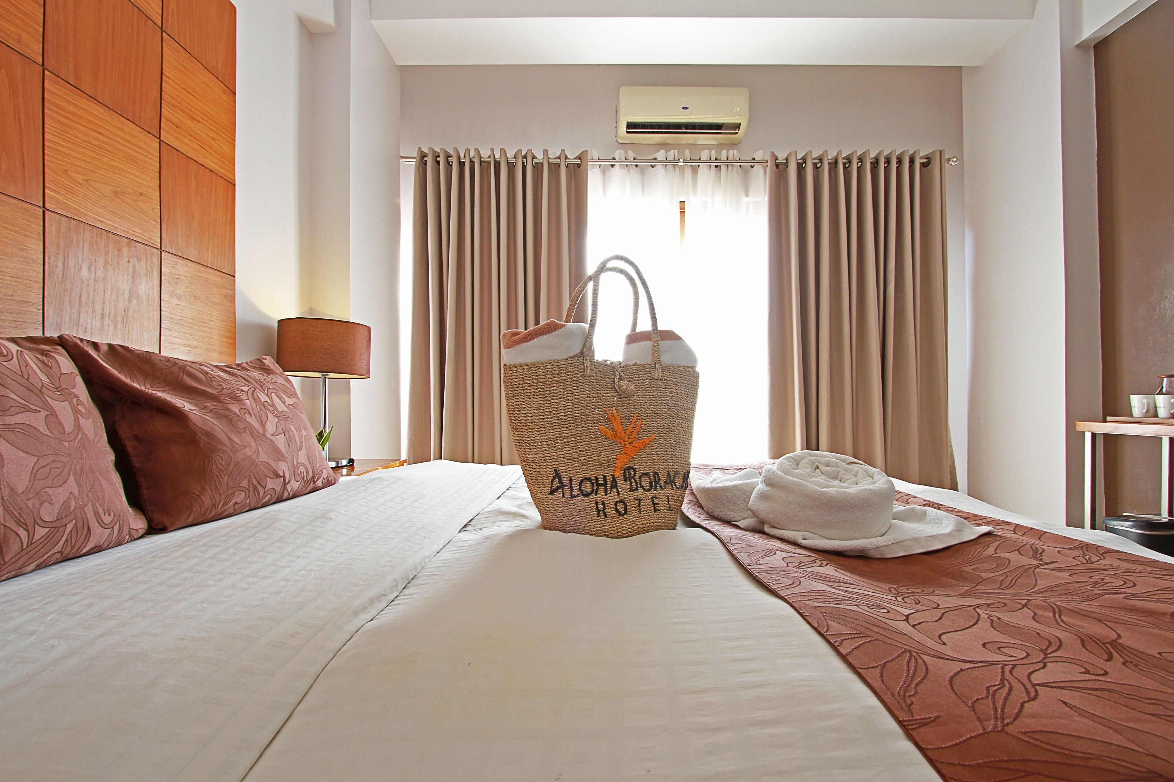 A hotel room with a bed, a headboard, a window with curtains, an air conditioner, and a decorative wall panel.